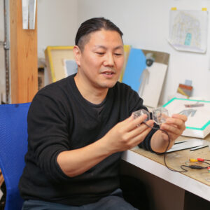 瀧健太郎さんの写真。作業台の椅子に座り、小さな装置を手に持っている。