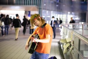 鮮やかなオレンジ色のTシャツを着て赤いバンダナを頭に巻き、ギターを弾いているひと