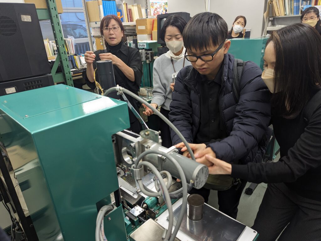 日本点字図書館にて、点図を製作する機械を森氏が触っている様子の写真。森氏の隣にいる女性が機械について説明し、その様子を女性３人が見守っている。