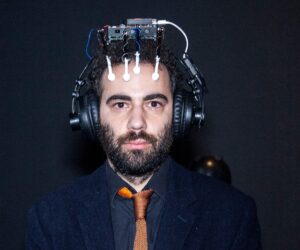 実験的な装置を頭に装着した男性の写真。スーツとネクタイを着用し、真剣な表情でカメラを見つめている。