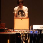 電子機器に囲まれ、演奏をしている男性の写真