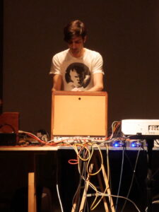 電子機器に囲まれ、演奏をしている男性の写真