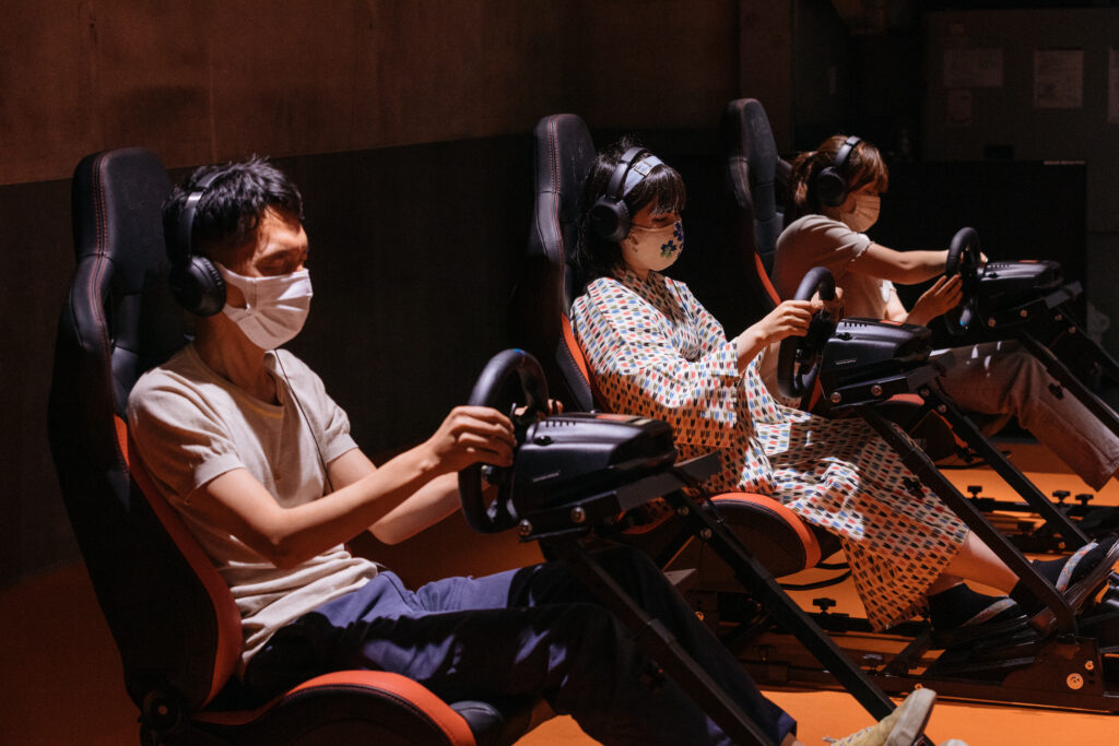 3人の男女がレーシングシミュレーターの座席に座り、ハンドルを握っている。全員がヘッドホンとマスクを着用しており、集中してシミュレーターを操作している様子。暗い背景に赤い座席が映えている。