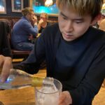 金髪の男性がバーのテーブルに座り、ボトルから氷入りのグラスに飲み物を注いでいる。彼は黒い服を着ており、集中してグラスを持っている。背景には別のテーブルで会話を楽しむ人々が見える。