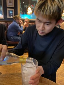 金髪の男性がバーのテーブルに座り、ボトルから氷入りのグラスに飲み物を注いでいる。彼は黒い服を着ており、集中してグラスを持っている。背景には別のテーブルで会話を楽しむ人々が見える。