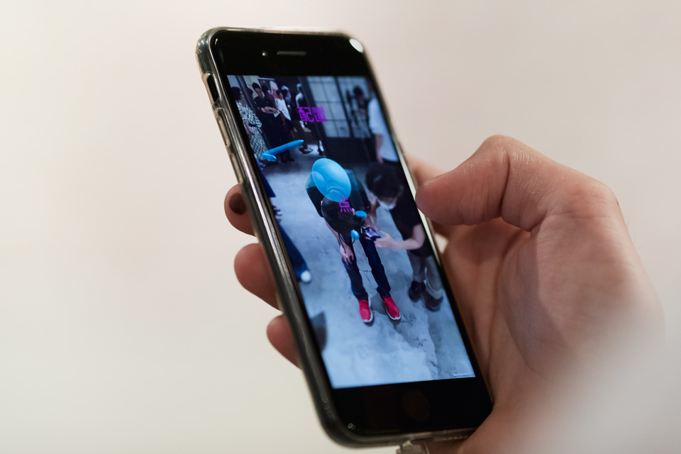 手に持ったスマートフォンの画面に、拡張現実（AR）技術を使っている様子が映し出されている。画面には青い球体とその背後に立つ人々が見える。スマートフォンを持つ手がしっかりと画面を支えている。

