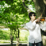 緑豊かな公園で、サングラスをかけた男性が木のそばでバイオリンを演奏している。男性は白いシャツとジーンズを着用しており、集中して楽器を弾いている様子。背景には明るい緑の木々が広がり、穏やかな雰囲気が漂っている。
