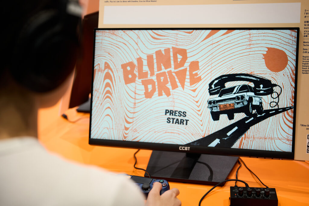 ヘッドホンを着けた人物がゲーム「Blind Drive」をプレイしている様子。画面には「Blind Drive」と「PRESS START」の文字が表示されており、車が電話を運転しているイラストが描かれている。背景はオレンジ色の模様になっている。