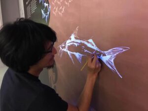 黒い髪の男性が、壁に白い魚の絵を描いている。男性はメガネをかけていて、絵に集中している。背景には魚のシルエットがいくつか見える。