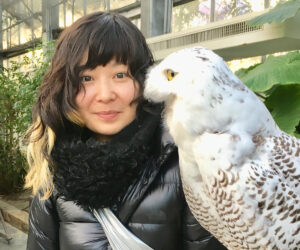 女性が大きな白いフクロウと一緒に写っている。女性は黒いコートを着て、微笑んでカメラを見ている。背景には緑の植物が見える。