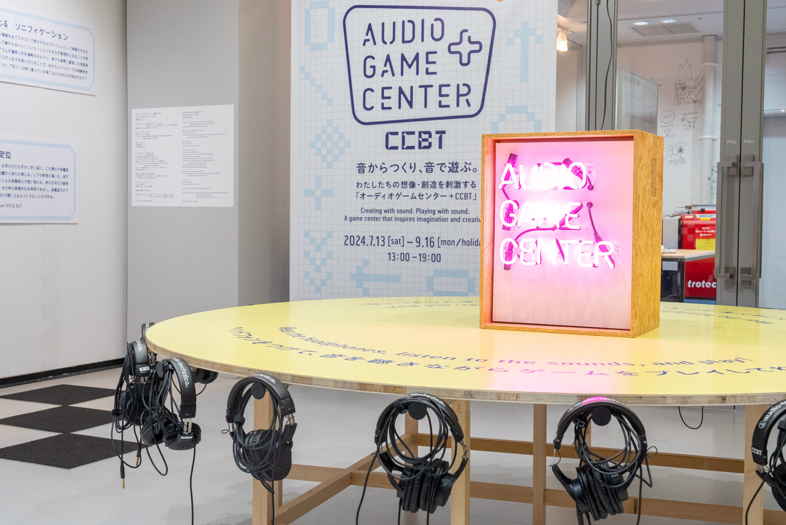 「AUDIO GAME CENTER」という文字が大きく表示されているサインと、周囲にヘッドホンが配置された円形のテーブルが写っています。背景には「CCBT」とイベントの開催日程が書かれたパネルが見えます。